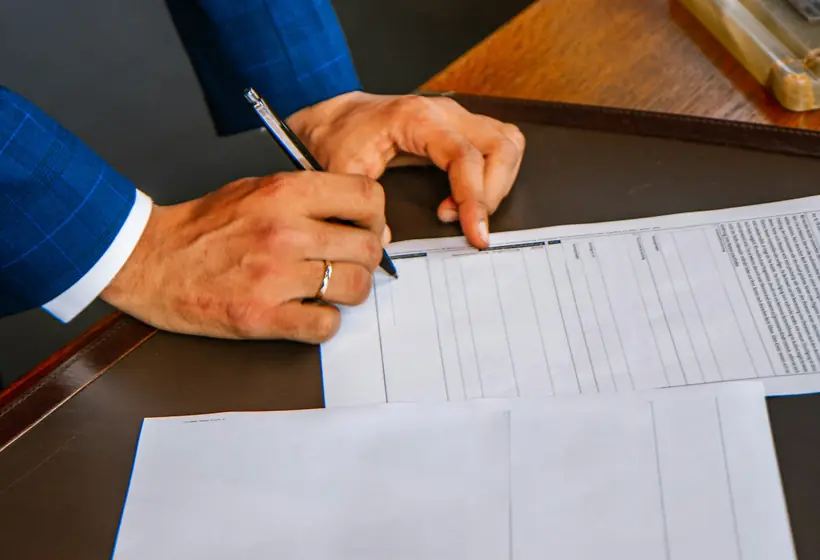 Person filling in a form at a desk with reading glasses beside it